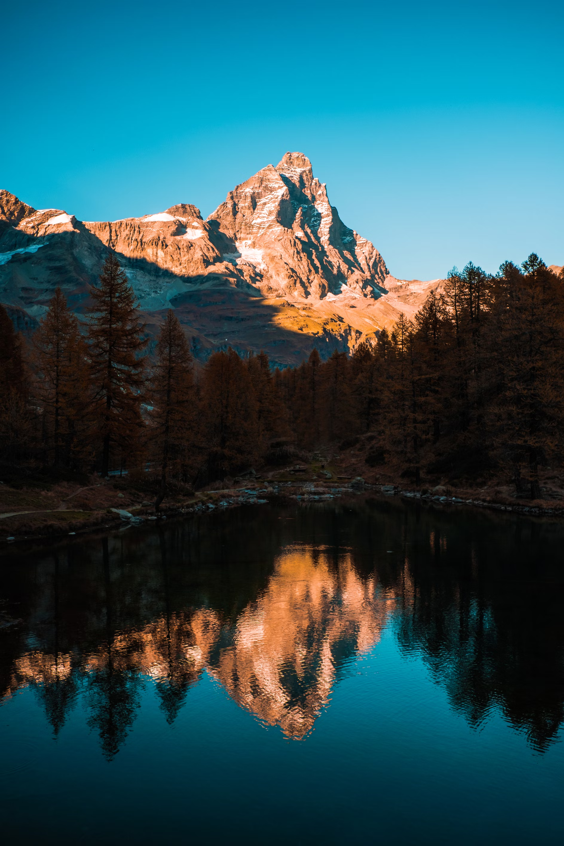 this is a landscape photo of a lake in the forest. the trees around the lake and a mountain range behind it are reflected on the surface of the lake. the photo is taken from the shore of the lake, with the water in the foreground. the trees are on the left and right of the photo. the mountain is in the background and is lit by the sun. the sky is blue and clear. the overall tone of the photo is peaceful and serene.