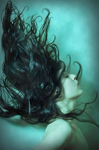 the image is a closeup portrait of a woman submerged in water. her dark hair swirls and dances around her, creating a sense of weightlessness and movement. the lighting is soft and diffused, giving the image a dreamlike quality. the overall palette of blues and greens contributes to the underwater theme and creates a sense of tranquility and peace.