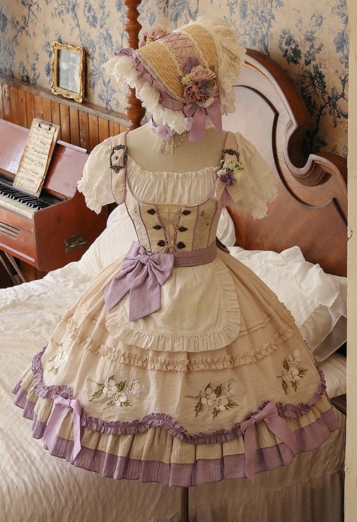 a full, vintagestyle lolita dress is displayed on a mannequin in an antique bedroom. the dress is a pale peach color with floral embroidery, lace trim, and purple ribbon accents. a matching bonnet with flowers is perched on the mannequin's head. the room features ornate wallpaper and a wooden headboard, adding to the vintage aesthetic. the scene is lit with natural light, creating a soft and inviting atmosphere.