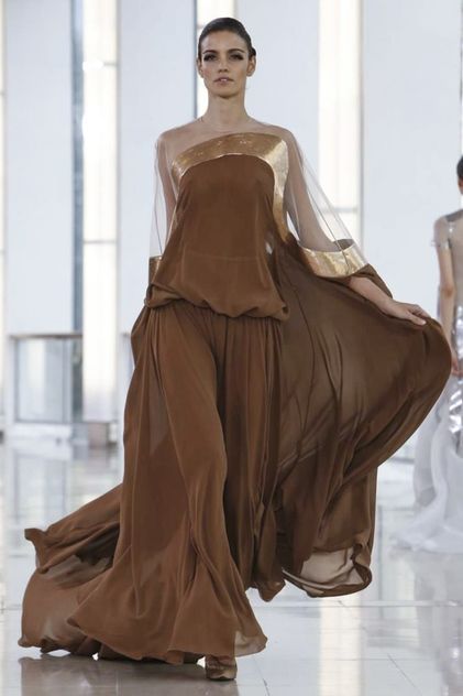 a fashion model walks down a runway in a long, flowing brown gown with a gold trim. the dress is very elegant and chic, and the model is showcasing it perfectly. the gown is made of a silky, chiffonlike fabric that drapes beautifully. the model is wearing simple makeup and her hair is styled in a sleek, straight style. she is walking gracefully down the runway, her pose is confident and beautiful. the dress is a perfect example of haute couture fashion.