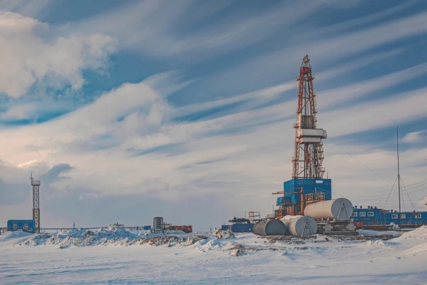 a large oil rig stands in a snowy, arctic landscape. the rig is composed of a tall tower and a complex network of pipes and machinery. the ground is covered in snow, and the sky is a mix of white and blue. the rig is a testament to the industrial activity that takes place in this remote region of the world.