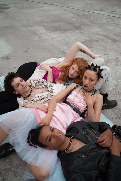 our young adults, a diverse group of friends, are lying together on a concrete ground. their fashion style is alternative and edgy; a mix of grunge, punk, and rock influences. their clothing includes pastel colors and dark colors, showing diverse styles and expressions. they appear relaxed and carefree, enjoying each other's company. the setting is outdoor, bathed in natural daylight. the photo is candid and captures a real moment of friendship among the group.