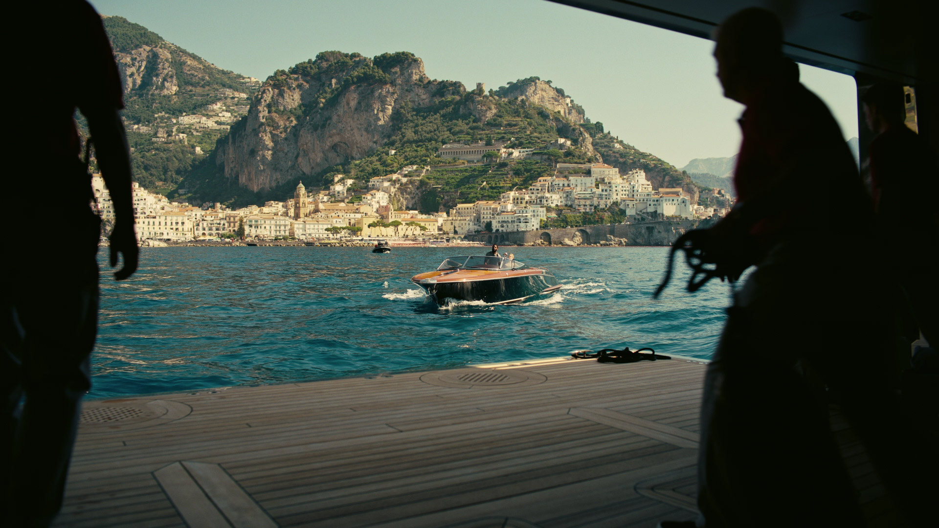 a view of a coastal town from a wooden dock. there is a small boat speeding away from the dock towards the town. two people in silhouette are standing on the left and right sides of the image. the town is nestled between green mountains and the blue water.