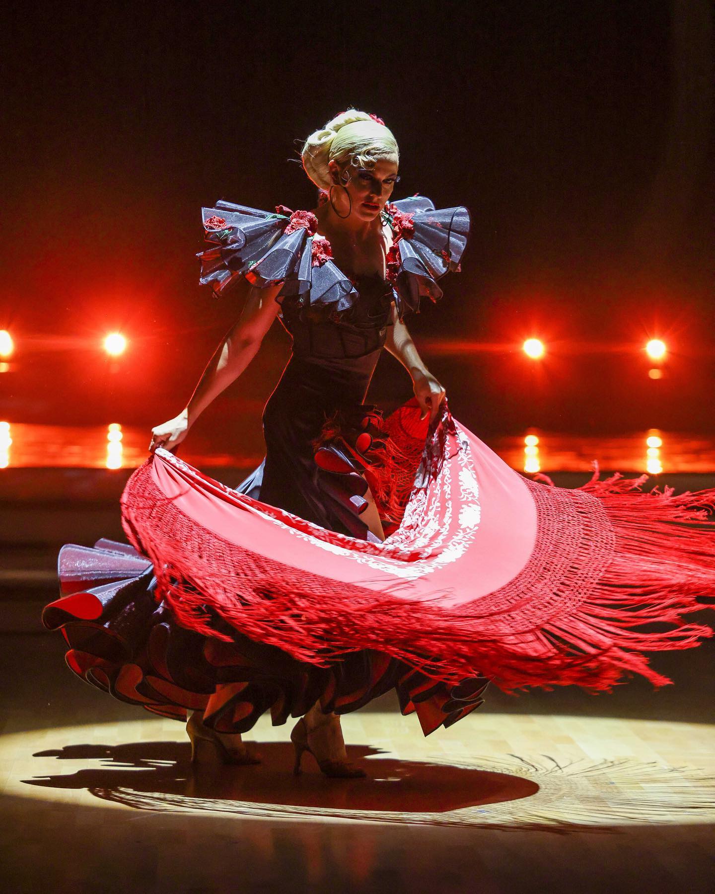 a female flamenco dancer in a black and red dress with ruffles is captured midperformance on a stage. she is moving gracefully, her dress swirling around her as she dances. the stage is lit with warm, dramatic lighting.
