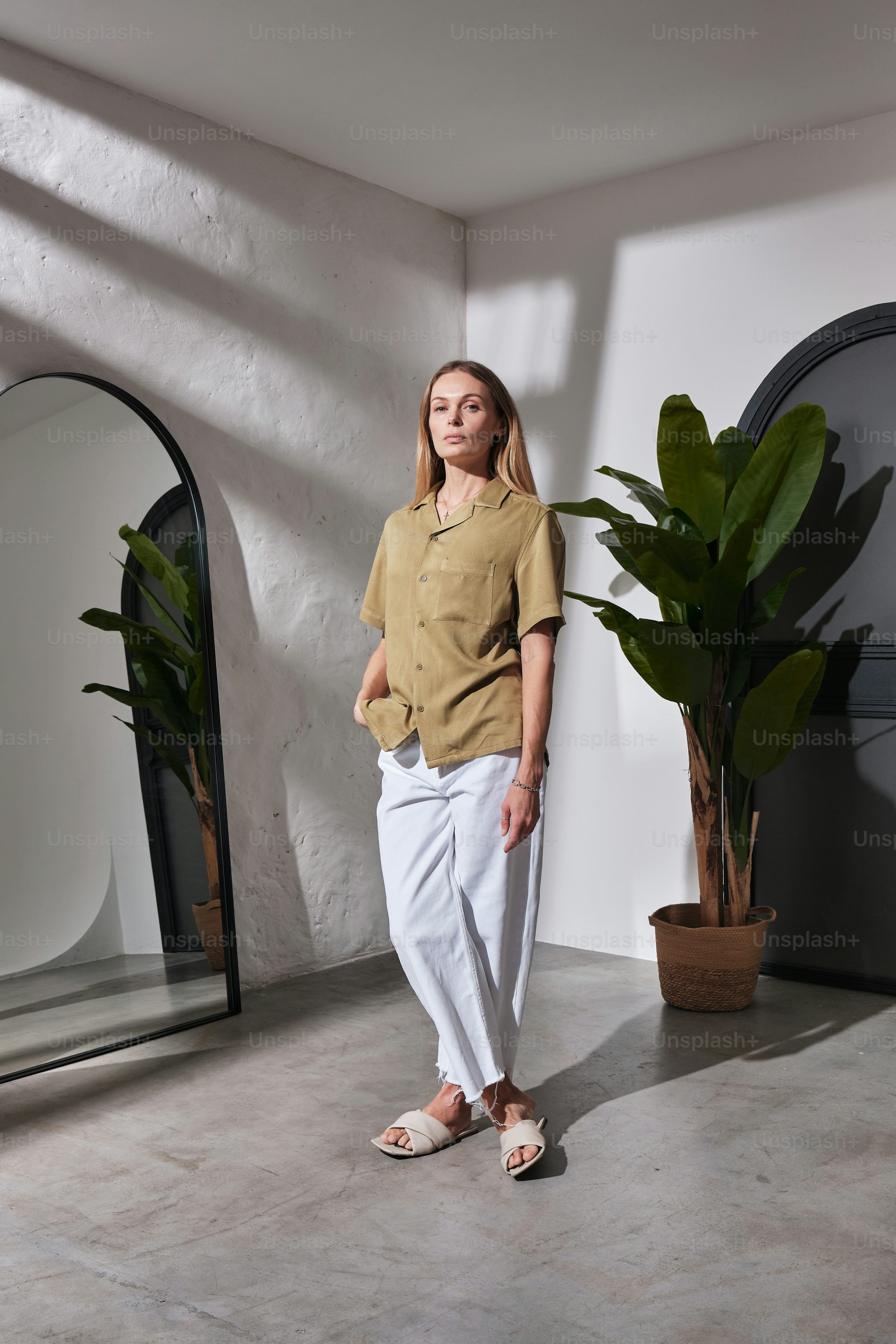 a woman stands in a minimalist room, bathed in sunlight. she wears a light beige shirt and white pants, and beige sandals.  she is positioned in front of a fulllength mirror reflecting a plant and parts of the room. a large potted plant stands behind her. the overall aesthetic is modern and serene.