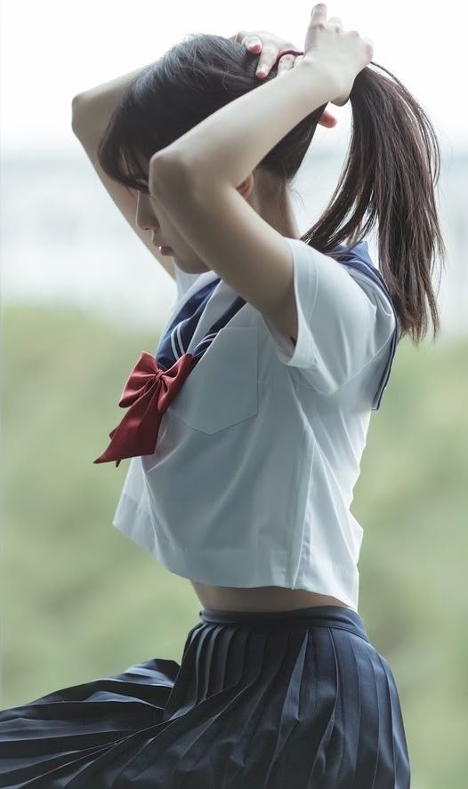 a girl in a japanese school uniform is tying her hair into a ponytail. she's wearing a white cropped shirt with a blue sailor collar and a red bow. she also has on a dark pleated skirt.