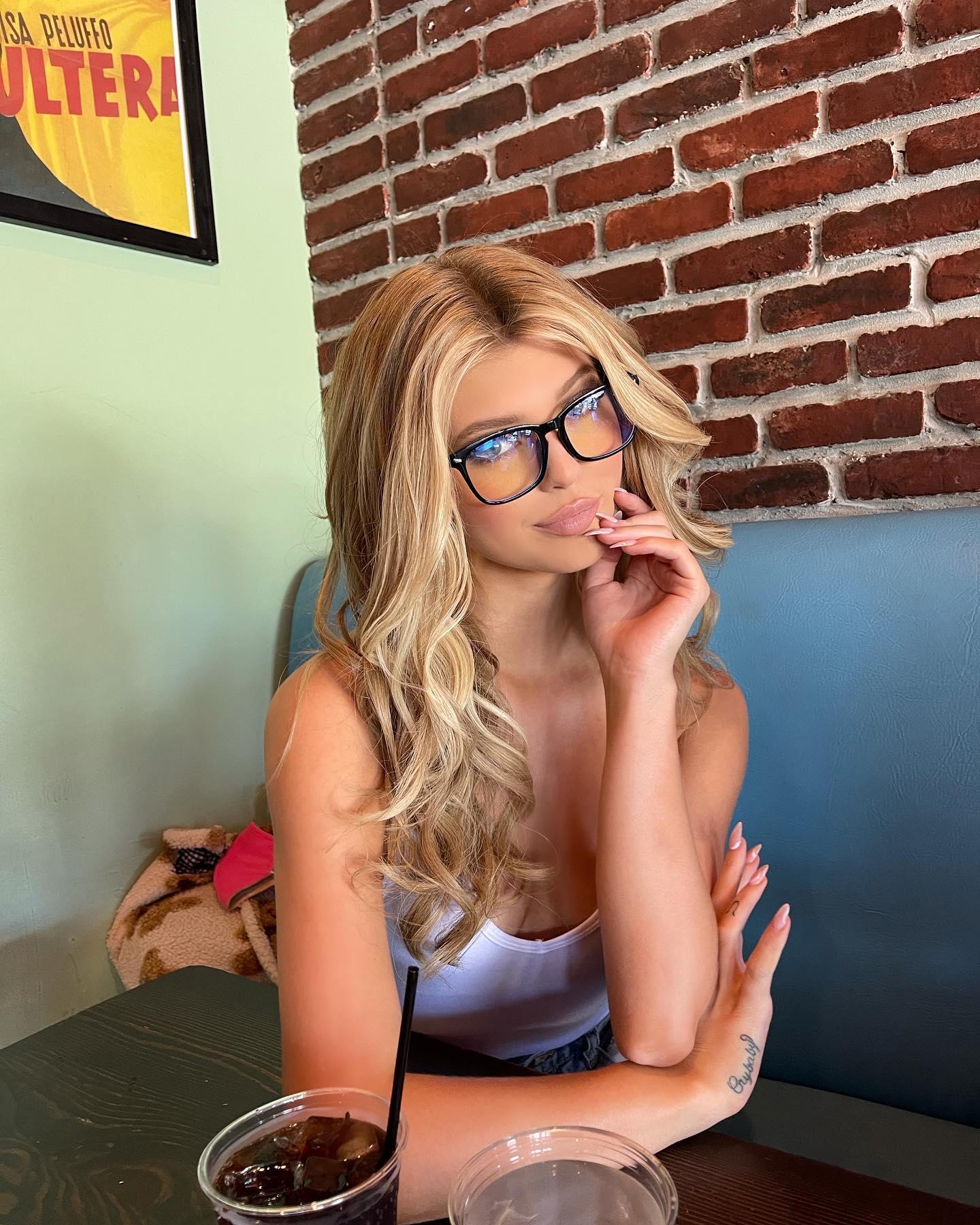 a woman with blond hair and glasses sitting at a table with two drinks in front of her. she is wearing a white top and has a tattoo on her arm. there is a brick wall behind her and a poster on the wall to the left.