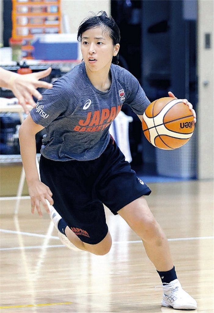 a young japanese female basketball player, wearing a grey japan basketball jersey and black shorts, is shown dribbling the ball during an indoor practice session. she is intently focused on the ball as she moves down the court, her expression one of determination.  the image captures her in motion, and her white sneakers are clearly visible. the background is blurred, with hints of court lines and some equipment visible.  the overall feeling is one of action and dedication to the sport.