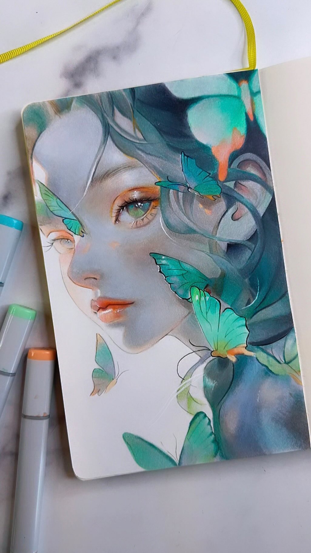 the image shows a closeup view of a watercolor painting or drawing on a sketchbook page. the artwork features a young woman's face, partially obscured and blended with soft, pastel colors, primarily teal and green. several butterflies in varying sizes and shades of these colors are scattered around her face and hair, creating a dreamy, ethereal atmosphere.  the style is delicate and feminine.  a set of alcoholbased markers is partially visible in the lower left corner, suggesting the medium used. the overall impression is one of beauty and tranquility.