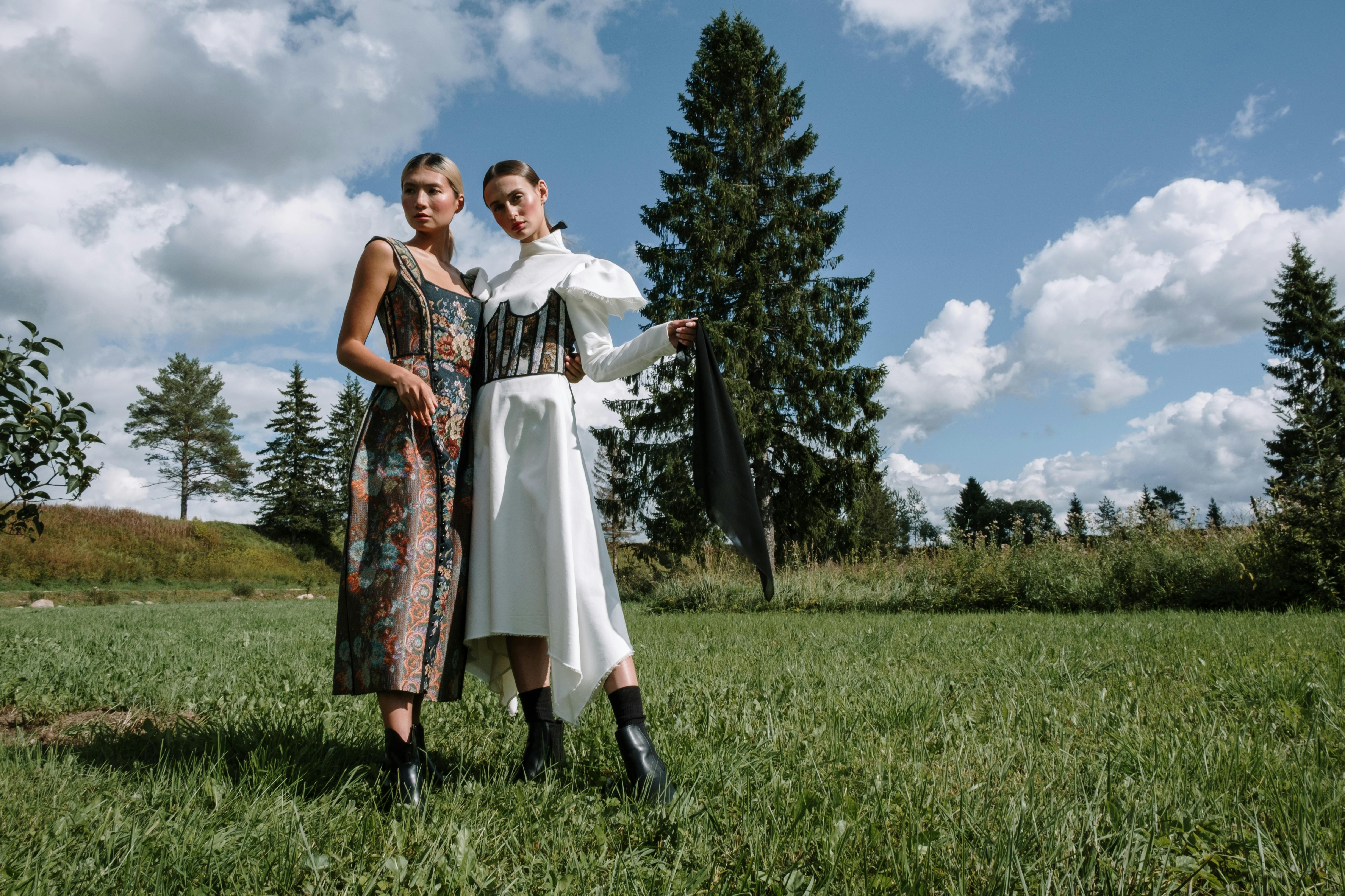 wo female models stand in a grassy field, posing for a fashion photoshoot.  the backdrop is a picturesque landscape of trees, a bright blue sky with fluffy clouds, and lush green grass. one model wears a long white dress with a black corset detail and the other wears a richly patterned, long floral dress.  both models wear black ankle boots. the overall mood is elegant and artistic. the photo is taken on a sunny summer day.