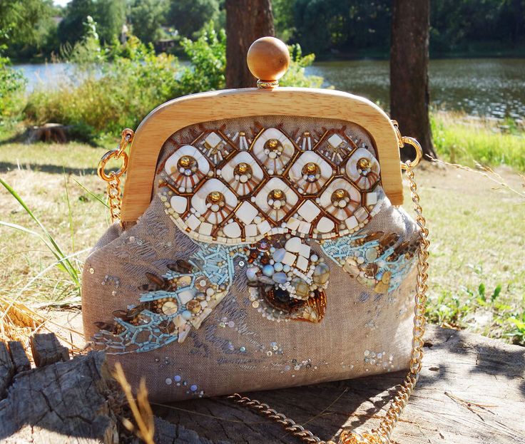 a beaded and embroidered purse with a wooden frame, sitting on a wooden surface in a nature scene. the purse is beige with gold embellishments, and has a gold chain strap. it is sitting on a wooden surface, with a lake and trees in the background. the leaves are turning yellow and orange, indicating it is autumn.