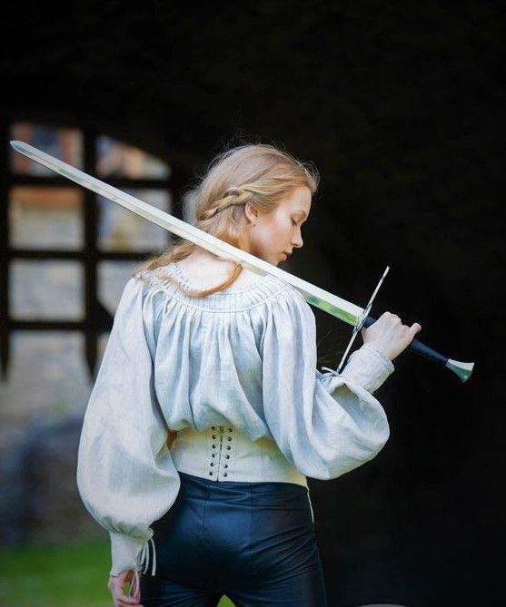 a young woman with long blonde hair is wearing a white shirt with a corset and black pants. she is holding a sword in her right hand, facing away from the camera. she is looking down at the sword. the sword is pointed down, and she is holding it with her right hand at the hilt, her left hand resting on the blade near the hilt. the background is dark, but there is a window with green plants visible.
