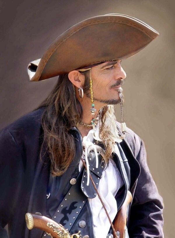 the image shows a man dressed as captain jack sparrow from the pirates of the caribbean franchise. he is wearing a large brown pirate hat, a black jacket, a white shirt, and a leather belt. he has long brown hair, a beard, and a mustache. he is wearing earrings and has beads in his hair. he is also holding a pistol. the background is a blurry brown color.