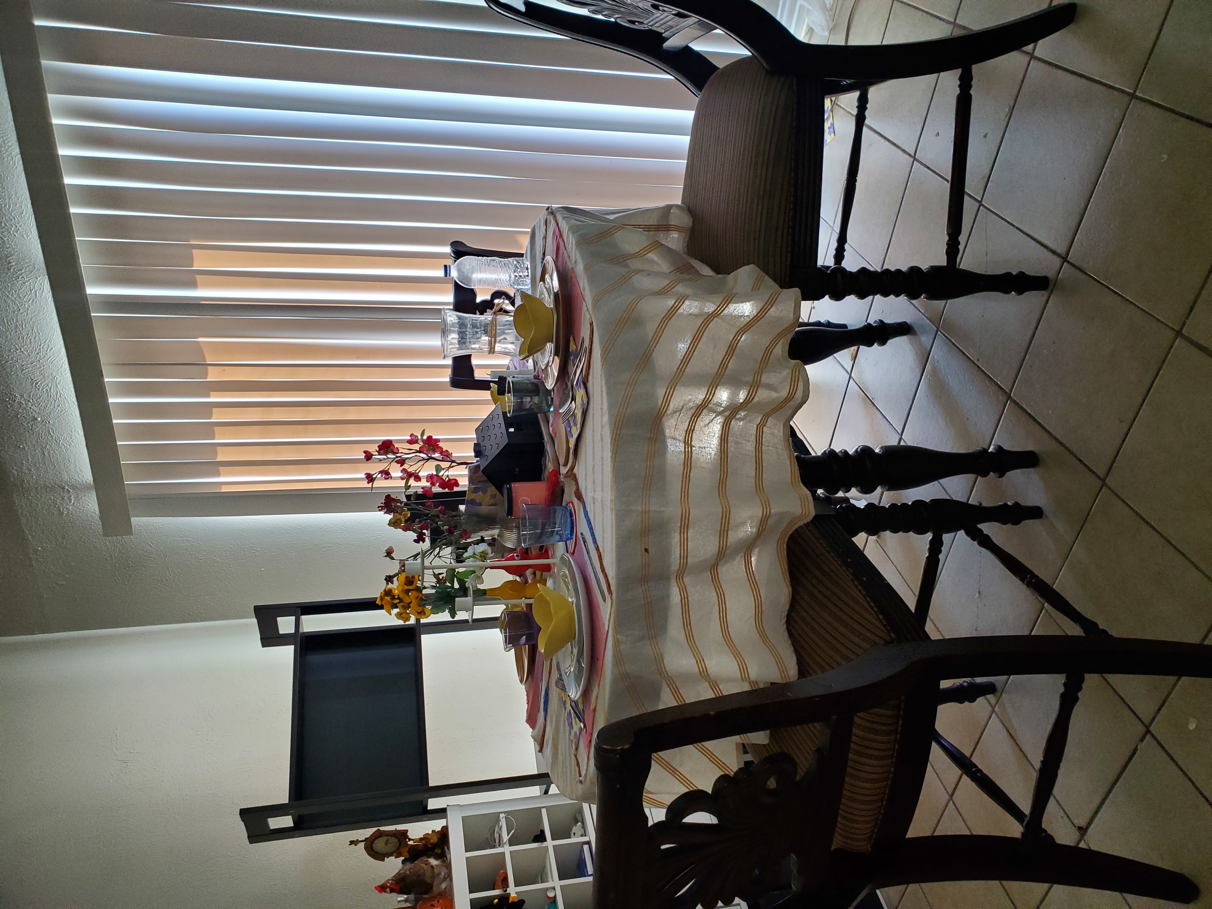 a dining table set for a meal, with a tablecloth, plates, cups, glasses, flowers, and decorations. the table is surrounded by chairs. in the background, there are blinds, tiles, a shelf, and decorations.