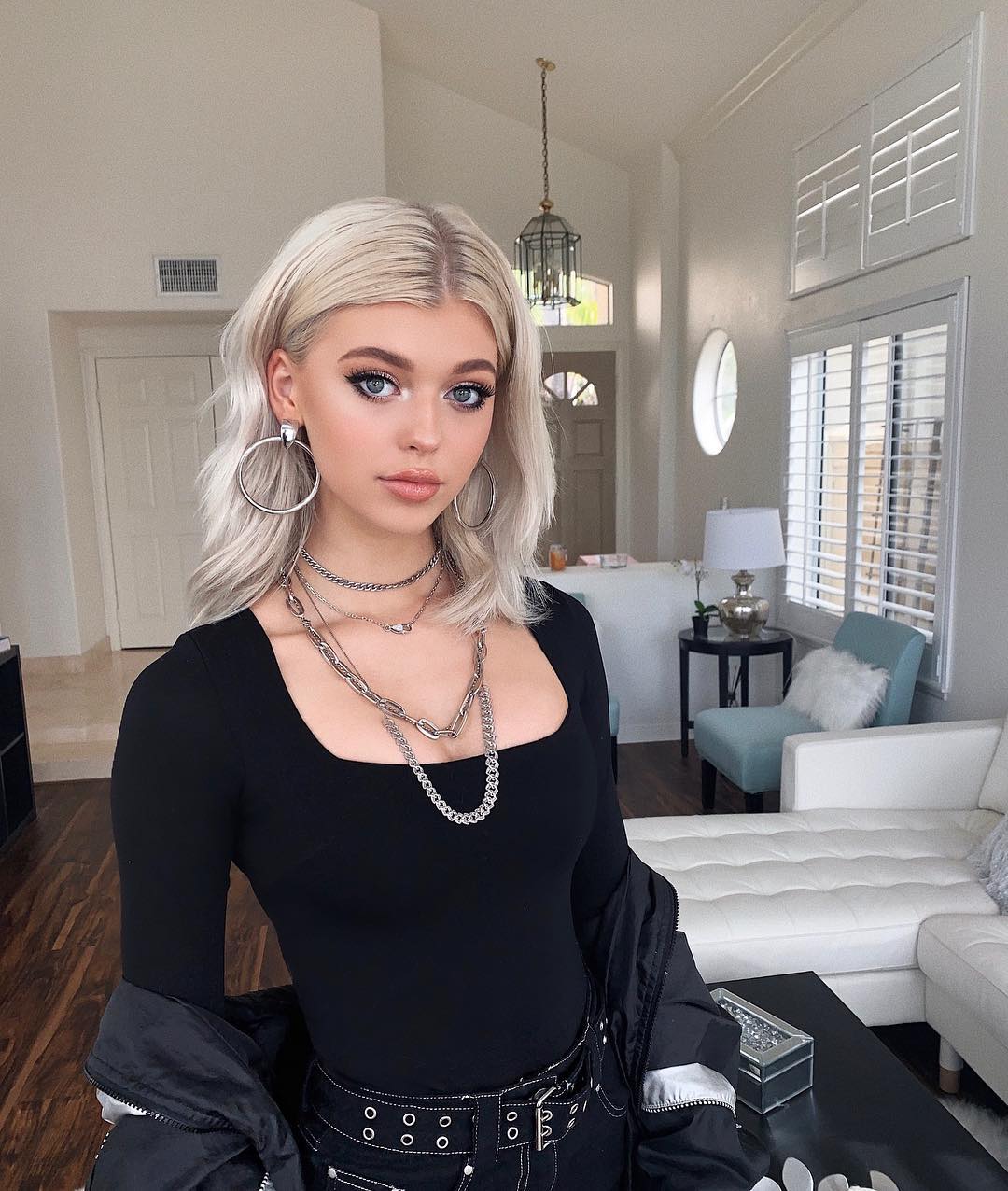 a young woman with blonde hair is posing in what looks like a living room. she's wearing a black top, a belt, and a jacket. she also has hoop earrings and multiple necklaces. the background includes a couch, a chair, a table, a lamp, a window with shutters, and a lighting fixture.