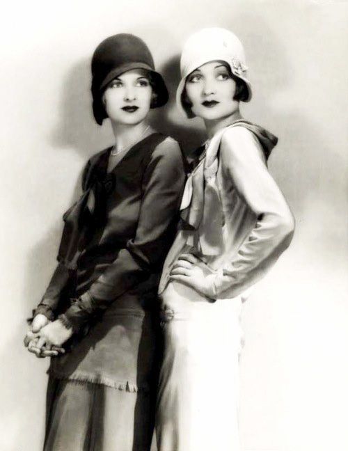 two women in 1920s flapper fashion pose for a studio portrait. one woman is wearing a black cloche hat, a dress with a frilled neckline, and a skirt with a fringed hem. the other woman is wearing a white hat with a black band, a blouse with a gathered neckline, and a white skirt with a gathered waistband. both women are looking at the camera.