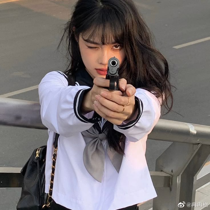 a young woman wearing a white school uniform with a gray bow and black accents is pointing a gun directly at the viewer. she is winking with one eye and has dark hair with bangs. she is wearing several rings. a black backpack with a gold chain is visible on her left shoulder. she appears to be standing next to a road behind a metal railing.