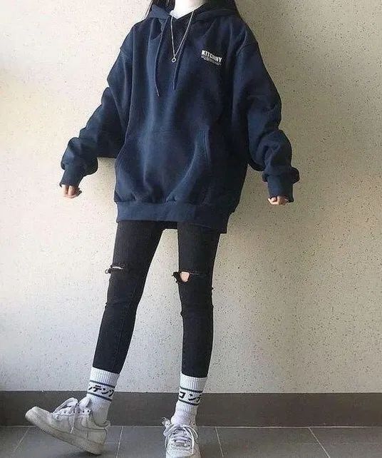 a woman in a dark blue hoodie with "kitchy" printed on it, wearing ripped black jeans, white socks with black lettering and white sneakers. she is standing on a gray tiled floor.