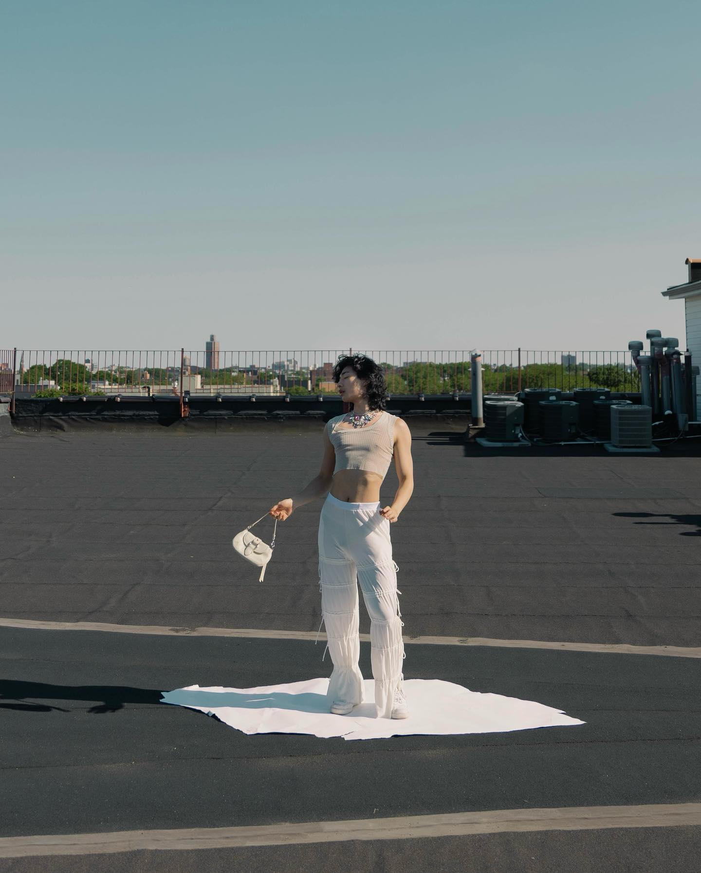 a stylish individual poses on a white sheet placed on a rooftop, bathed in sunlight. they wear a beige crop top and white pants with a playful fringe detail. they carry a small white purse and gaze away from the camera. the urban landscape provides a backdrop to this fashionforward moment.