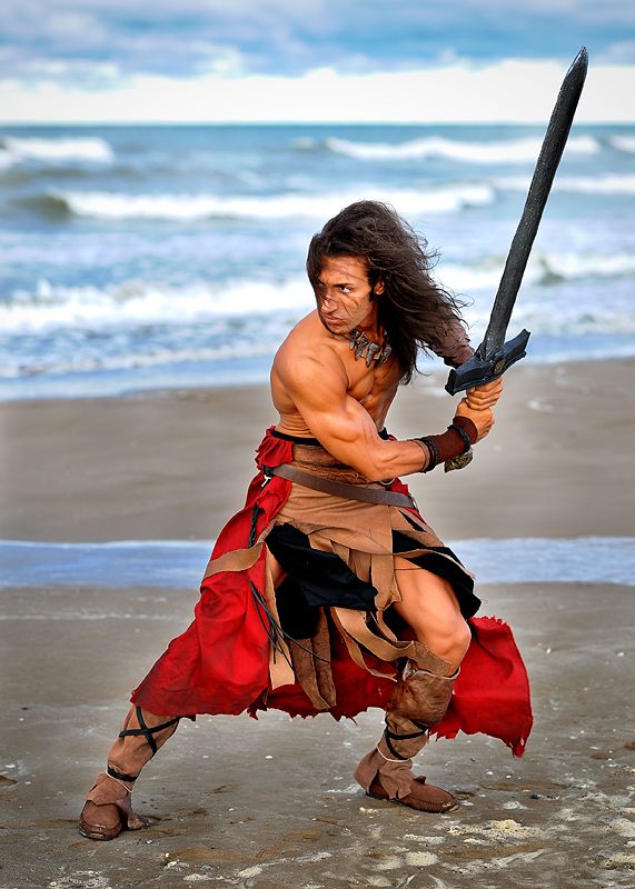 a man dressed as a barbarian warrior stands on a sandy beach. he is holding a sword in his right hand and his left hand is on the hilt. he is wearing a red and brown leather costume. the beach is in the background, with the sea in the distance.