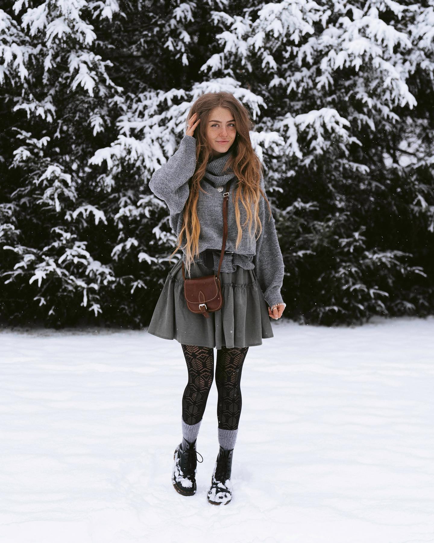 a young woman with long, light brown hair stands in a snowy landscape. she is wearing a grey oversized sweater, a grey pleated mini skirt, black patterned tights, and black doc martensstyle boots. she carries a small brown crossbody bag. the background features snowcovered evergreen trees. the overall style is casual yet stylish, with a touch of boho chic. the image evokes a feeling of winter and cozy outdoor fashion.
