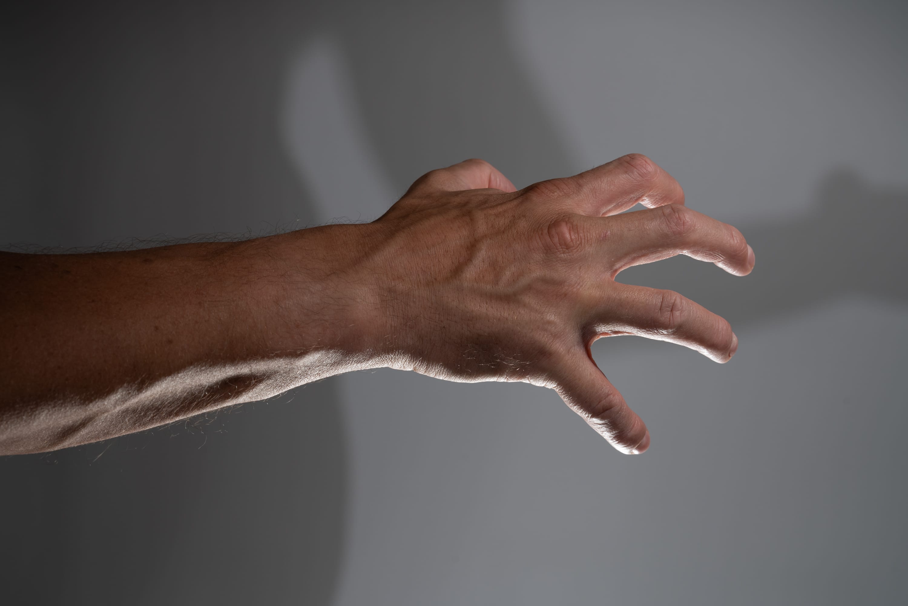 a closeup image featuring a man's hand and arm against a gray background. the hand is partially clenched, with fingers extended and separated. a distinct shadow of the hand and part of the body is projected onto the background, adding depth and visual interest. the lighting emphasizes the texture and veins of the hand, highlighting its natural contours and details.