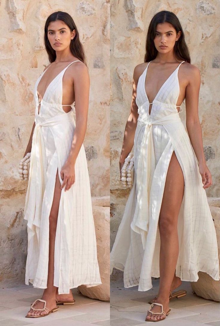 two images of the same model wearing a beautiful offwhite linen maxi dress with a high slit and a vneckline. the dress is tied at the waist and features a relaxed, flowing silhouette.  the model is wearing flat beige sandals and carries a small beaded clutch. the background is a rustic stone wall, creating a stylish and summery atmosphere. the images are nearly identical, showcasing the dress from slightly different angles.