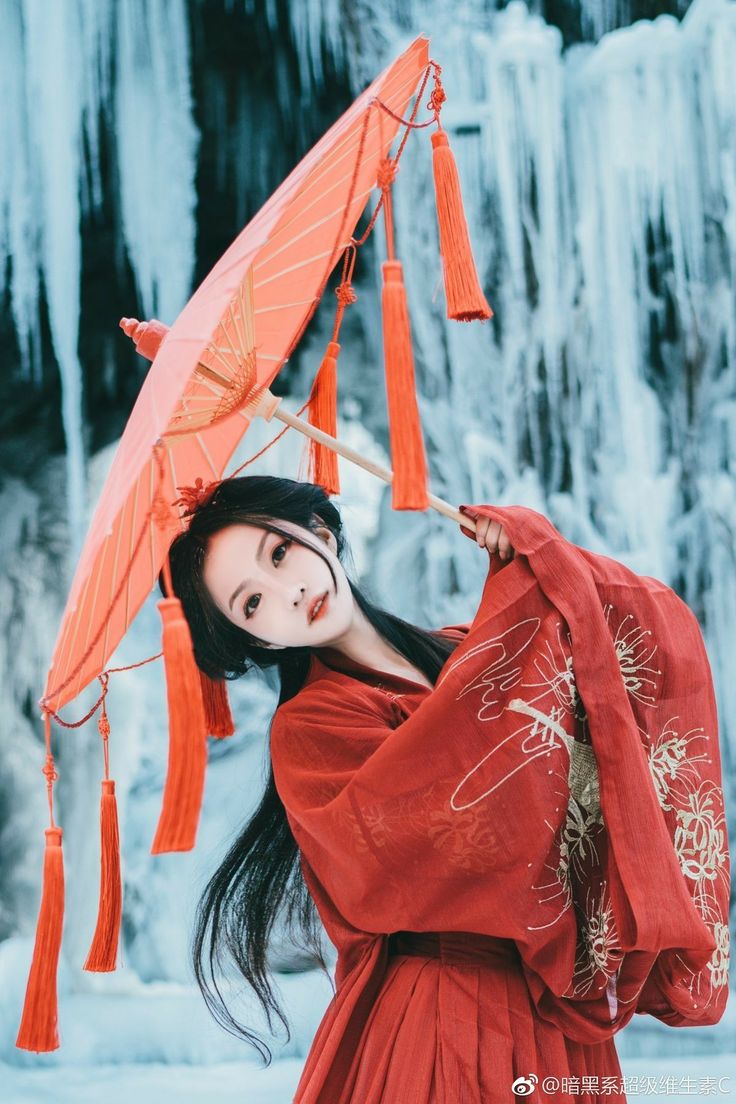 a young woman is wearing a beautiful red traditional chinese dress and holding a red umbrella. she is standing in front of a snowy background. her black hair is flowing, and she has a delicate and elegant look. the woman looks graceful and stunning in her traditional clothes.