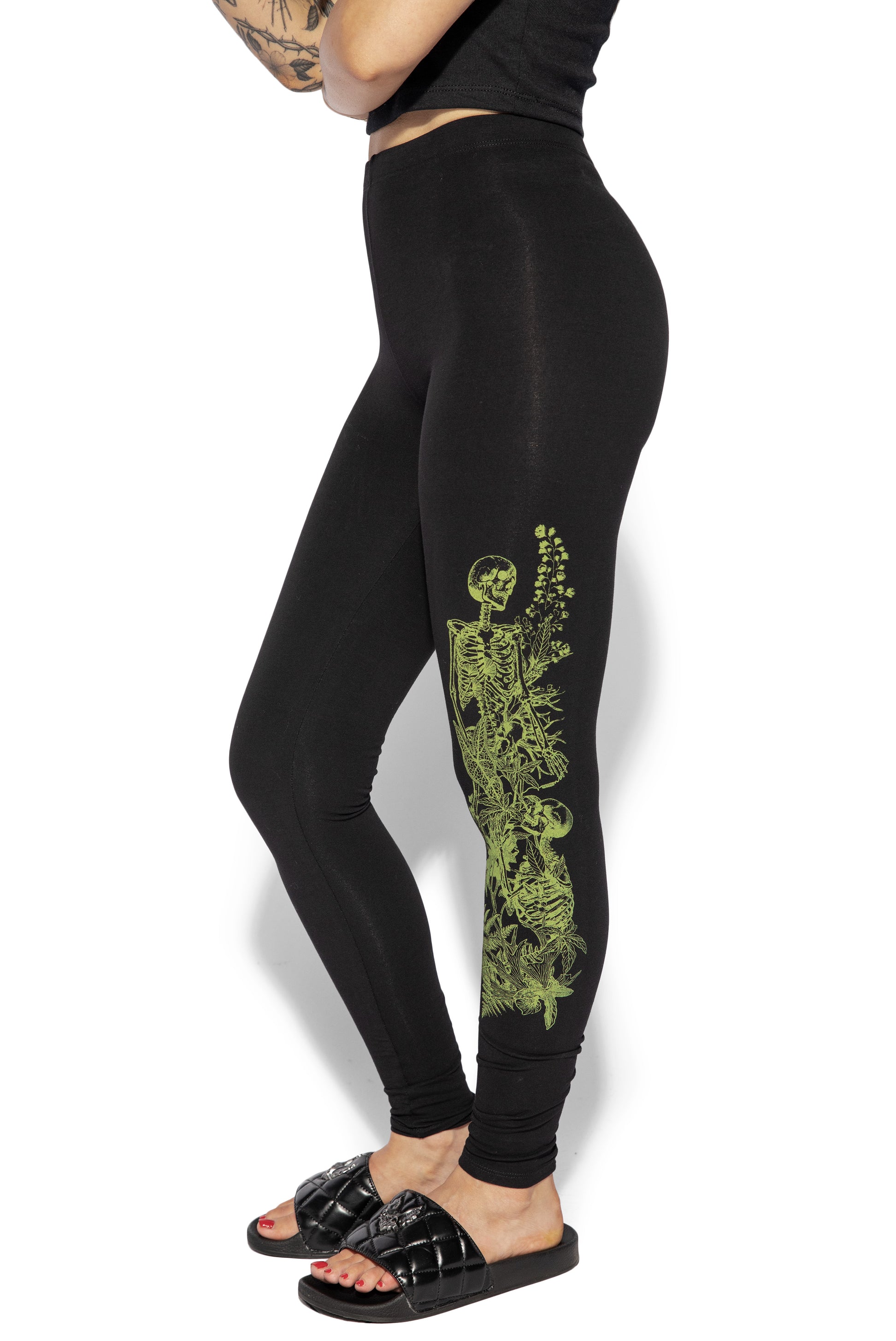 a side profile, fulllength studio shot of a model wearing black highwaisted leggings with a lime green skeleton and floral print on the side. the model is wearing black quilted slides and has tattoos on her arms. the leggings are casual and comfortablelooking.