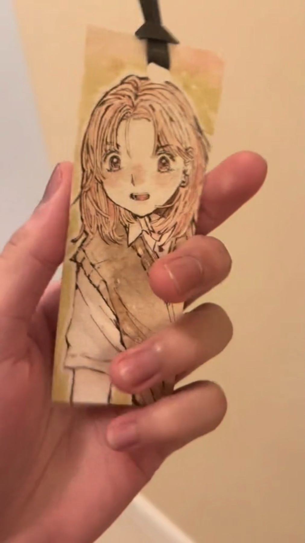 a hand is holding a bookmark with a cute anime girl drawn on it. the girl is smiling and has long hair, and she is wearing a school uniform. the bookmark is surrounded by a yellow background, and the girl's face is illuminated by a soft light.