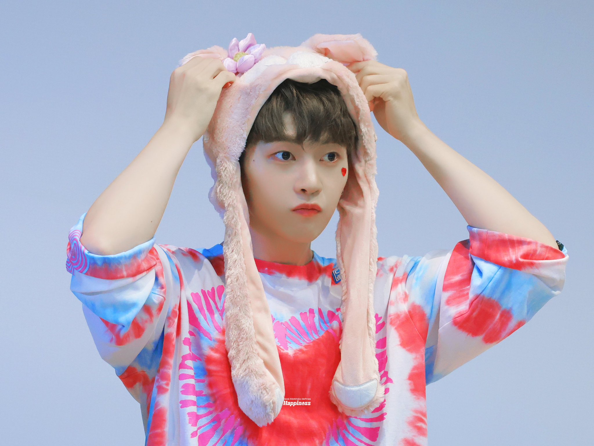 a young man with brown hair and a heartshaped sticker on his cheek is adjusting a pink, fluffy bunny ear hat on his head. he is wearing a pink and blue tiedye shirt with a heart design. the background is a light blue.