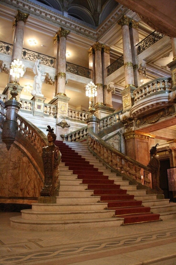 he image shows a grand, ornate staircase in a luxurious building. the staircase is made of marble and features intricate carvings and gold detailing.  there are marble columns flanking the stairs, and a red carpet runs up the center.  the architecture is opulent and suggests a palace or grand mansion.  statues and elaborate chandeliers are visible, adding to the overall sense of grandeur and luxury. the multiple levels of balconies and railings suggest a multistory building of significant size and importance.  the lighting is soft and warm, highlighting the details of the architecture and creating a dramatic and elegant atmosphere.