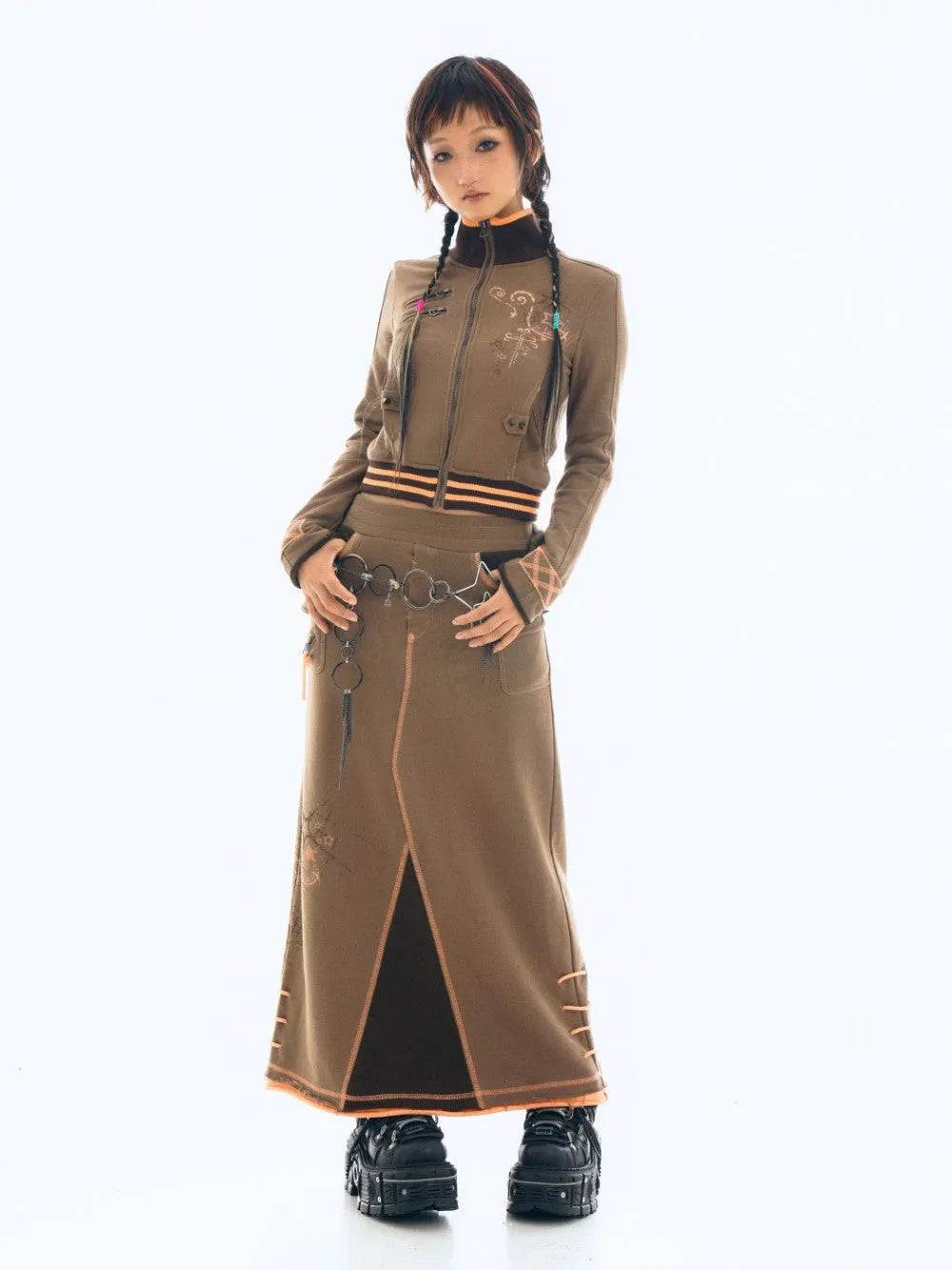 a full shot of a young woman wearing a stylish outfit. she is wearing a brown zipup jacket with orange details and embroidery, paired with a long brown skirt that has a center slit and orange stitching.  the skirt and jacket appear to be made of a soft, possibly fleecelike, material. she's accessorized with a belt and chunky platform boots. the model has dark hair styled in pigtails, and her pose is slightly angled, looking directly at the camera. the background is plain white. the overall style seems to be a blend of streetwear and japanese fashion influences.