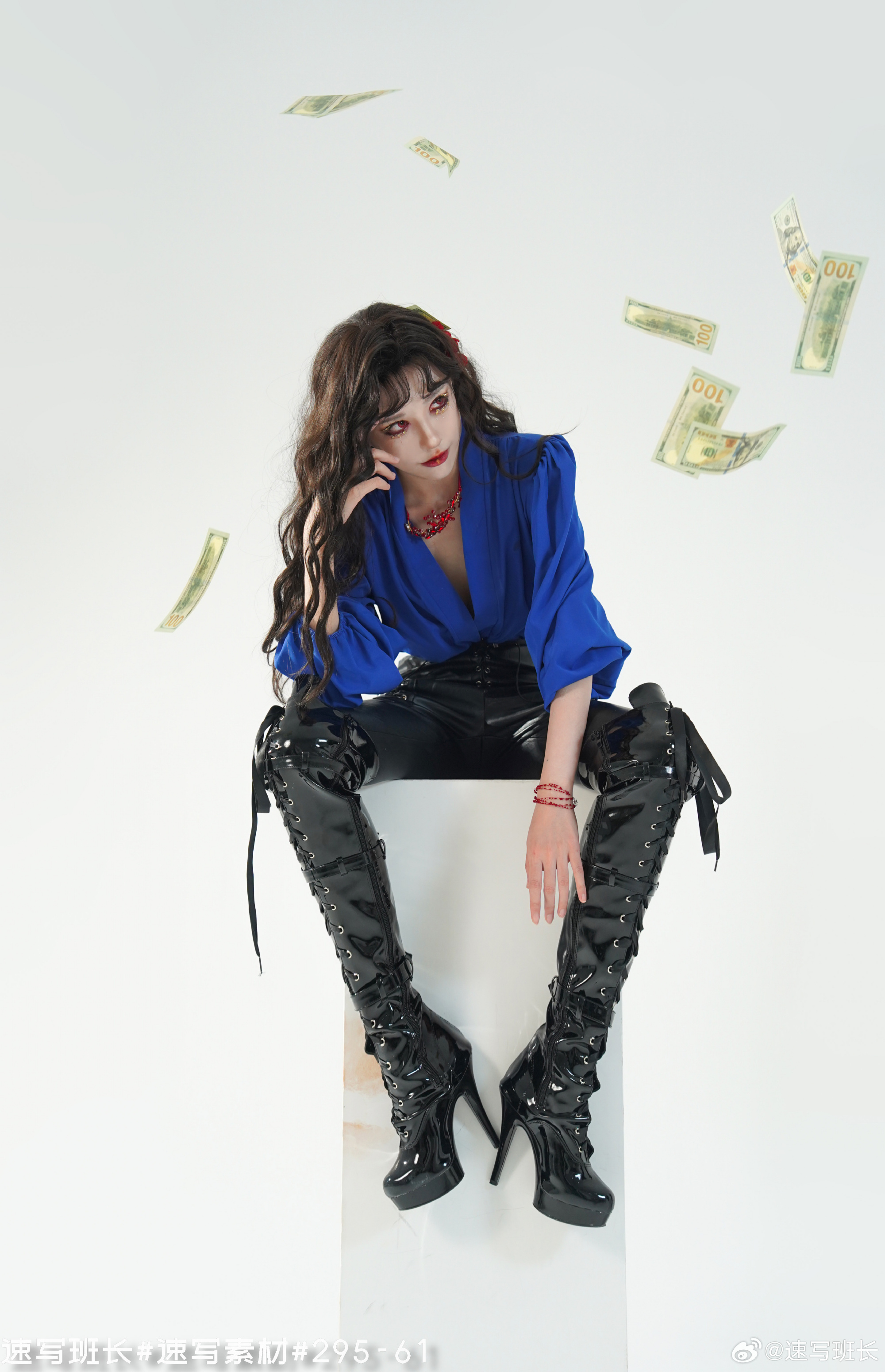a woman with long black hair and red lipstick is sitting on a white cube. she is wearing a blue long sleeve shirt, black leather pants, and black highheeled boots. she is looking down and to the left, and there are several us dollar bills floating around her. the background is white.