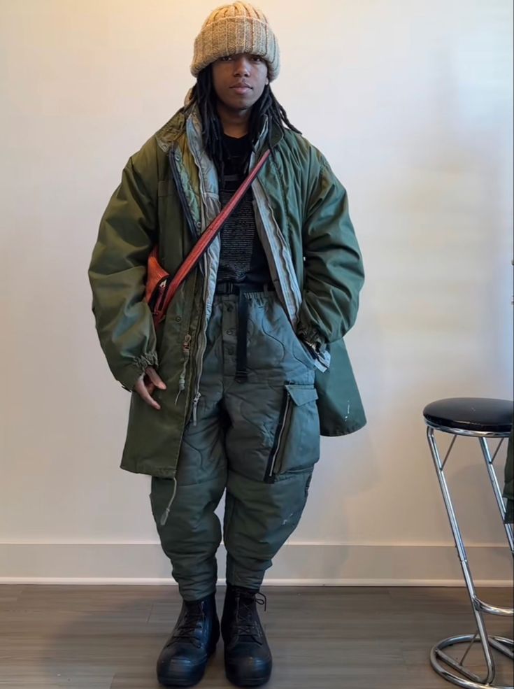 a full shot of a young person modeling an outfit. they are wearing an olive green long parka, olive green quilted cargo pants, black combat boots, a beige knit beanie, and a long orangered strap across their chest. the model has long, dark hair and is standing against a light wall with a metal stool in the background. the overall style is streetwear with a focus on layering and utilitarian elements.