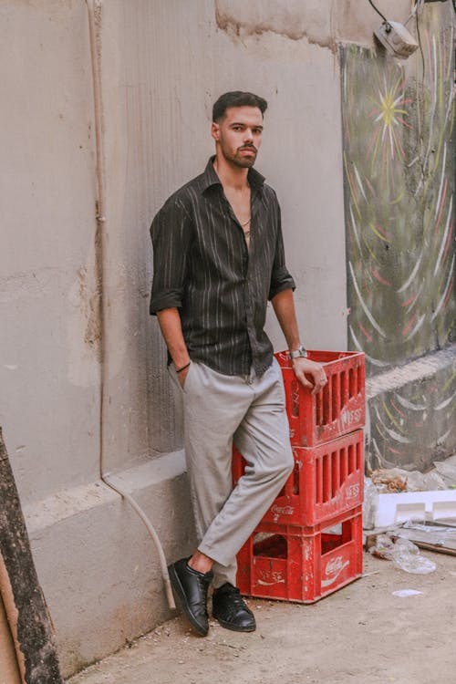the image shows a young, stylish man standing against a weathered, urban wall with graffiti visible in the background. he's dressed in a black and grey striped longsleeved shirt, light grey pants, and black sneakers. he is leaning against a stack of red plastic crates, likely used for beverages (cocacola branding is visible).  his posture is relaxed but his expression is serious. the overall setting is gritty and urban. the lighting suggests it's likely daytime.  the photograph's style is somewhat moody and slightly edgy.
