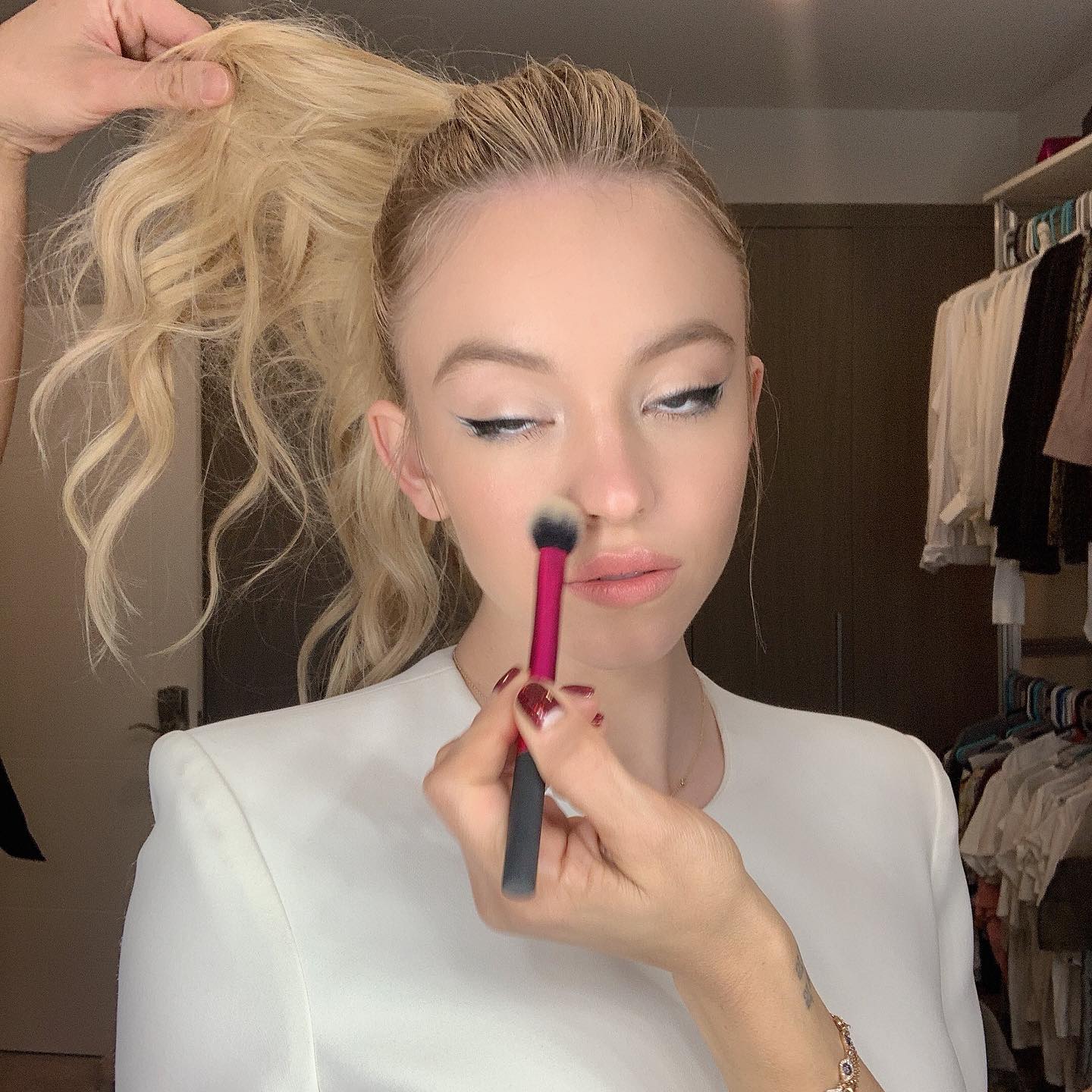 a young blonde woman with her hair in a high ponytail is having her makeup applied by a makeup artist.  the focus is on the application of blush to her cheeks using a small, angled brush. the woman is wearing a white blazer. the background shows a closet with clothes hanging inside, suggesting a gettingready scene, perhaps backstage at a photoshoot or for a video.  the overall mood is one of professional preparation and beauty.