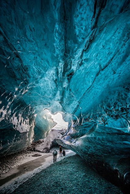 this is a stunning photograph of an ice cave interior. the cave's ceiling and walls are a brilliant, translucent blue, with intricate patterns and formations carved by the movement of the glacier. three figures stand near the entrance, providing scale and hinting at exploration. the cave opens to a bright, snowy landscape. the ground is covered in dark pebbles, and a stream of meltwater runs through the center. the overall scene evokes a sense of wonder and the raw beauty of nature.