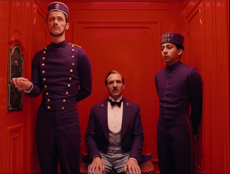 three men are in an elevator. two of the men are wearing purple uniforms. one man is sitting down in a chair while two men stand. the man sitting has a gold hat on his head and a black bow tie. the two men standing are both wearing purple hats and have gold lettering on them. the elevator is red with gold trim.  the man standing to the left is pressing the button in the elevator. the man standing to the right is looking straight ahead at the camera.  the man sitting is looking at the man standing to the right.  the elevator is very brightly lit.  the man in the chair is sitting on a chair with a gold and red pattern on it.  the elevator is very clean and wellmaintained.
