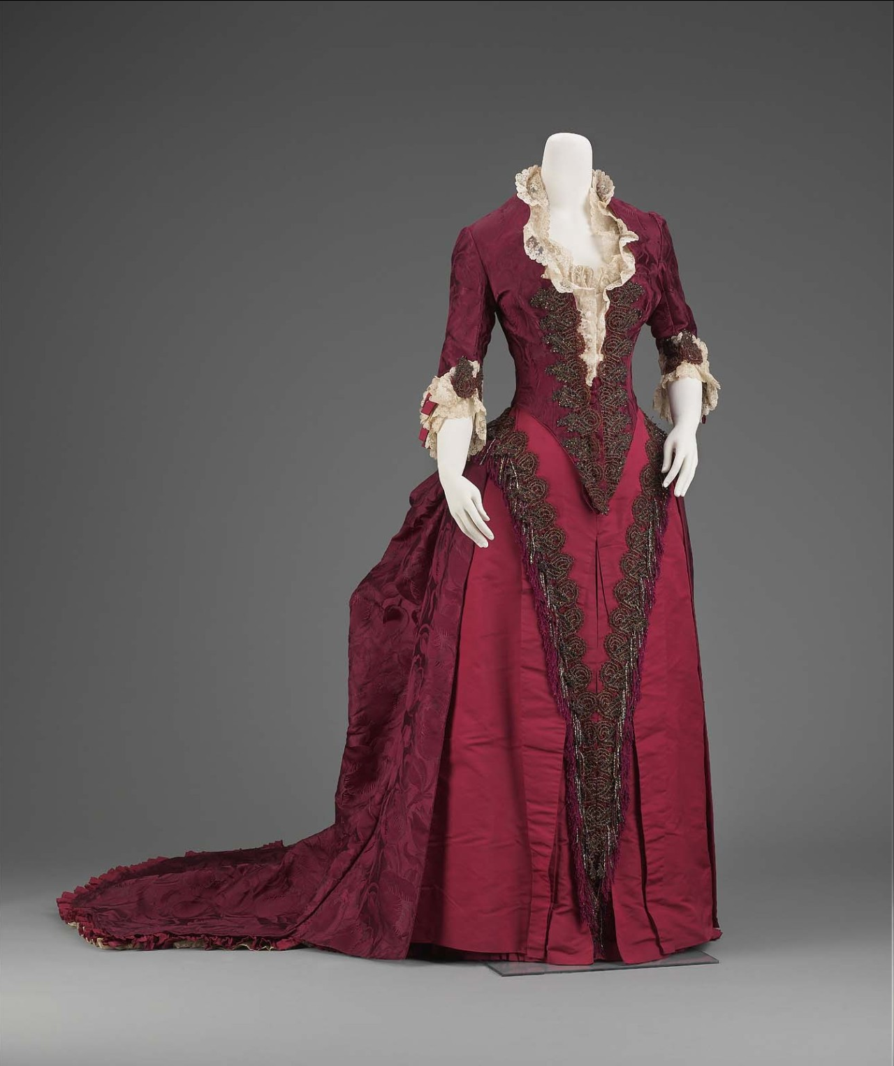 a fulllength view of a burgundy victorian era dress on a mannequin against a gray background. the dress features elaborate embroidery, lace detailing, and a long train.
