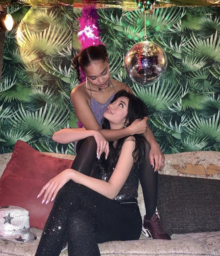 two young women are sitting on a couch, a black woman with a pink feathered headpiece and an asian woman in a black sequined outfit. they're smiling and laughing together as they look at each other. there is a disco ball hanging from the ceiling above them and palm leaves wallpaper behind. it looks like they're having a fun night out together.