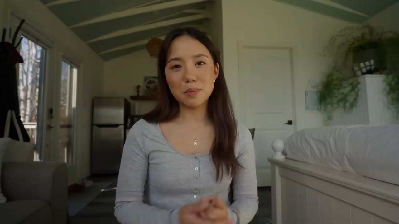a young woman with long brown hair is sitting in her living room and talking to the camera. she is wearing a gray shirt and has a necklace on. the room is bright and airy, with white walls and a blue ceiling. there is a couch behind her and a bed to her right. there is a door in the background.