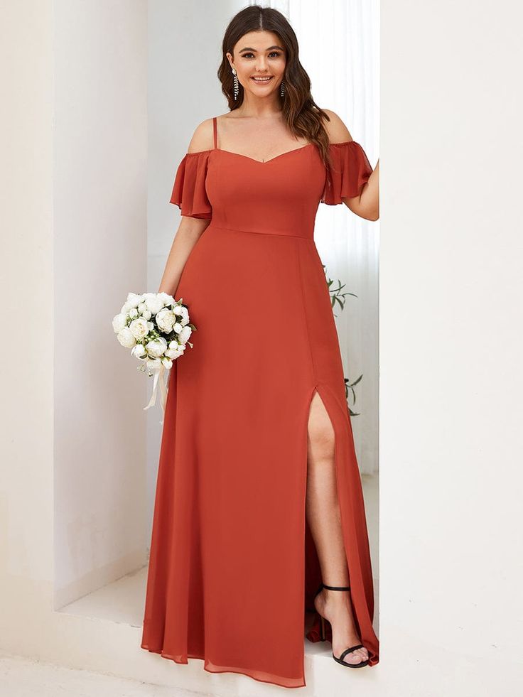 a woman in a beautiful red maxi dress, with ruffle straps and a slit, stands in front of a white wall. she is holding a bouquet of white flowers and is smiling at the camera.  she is wearing black heels.  she is ready for a wedding or a special occasion.