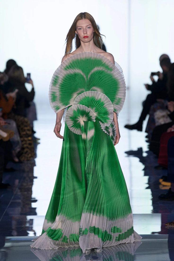 a woman model walks down the runway in a green and white pleated dress, which features a layered fanshaped top. the dress is cinched at the waist and flows down to the floor in a graceful skirt. the model has long blonde hair and simple makeup. she is wearing a green dress that is pleated and has a white trim at the bottom. the dress is very long and flowing and the model is wearing high heels. the background is a white wall and there are other people in the background. the overall aesthetic of the image is very stylish and elegant.