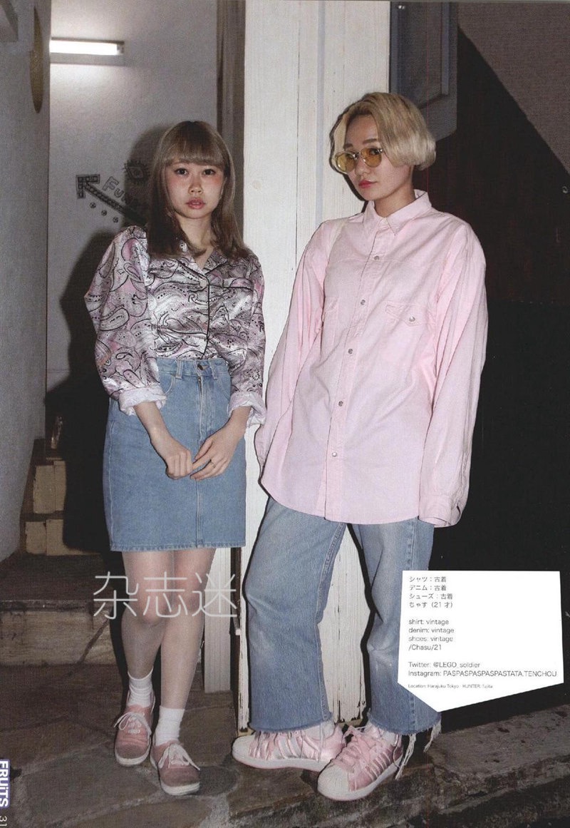 two young women pose for a street fashion photograph in japan.  the woman on the left has blonde bangs and wears a patterned shirt, denim skirt, white socks, and pink sneakers. the woman on the right has short, bleached blonde hair and wears an oversized pink buttonup shirt, baggy jeans, and pink and white sneakers. the background features a worn doorway with japanese text. the image has a slightly grainy, vintage aesthetic.
