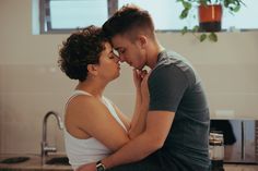 a man and a woman are embracing in a kitchen. the woman is wearing a white tank top and the man is wearing a blue tshirt. they are standing close together and looking into each other's eyes. there is a window in the background and plants are visible on the window sill. the kitchen is bright and airy, with white walls and a stainless steel sink. the lighting is soft and natural, creating a warm and intimate atmosphere.