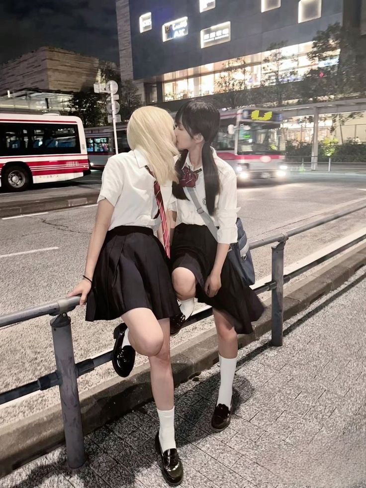 the image shows two japanese schoolgirls in uniform kissing on a city street at night.  they are wearing black pleated skirts, white blouses, and ties.  both girls are wearing white socks and black loafers. one girl has blonde hair and the other has dark hair. they are leaning against a metal railing, with buses visible in the background. the overall atmosphere is romantic and intimate, suggesting a young lesbian couple. the setting appears to be an urban area in japan.