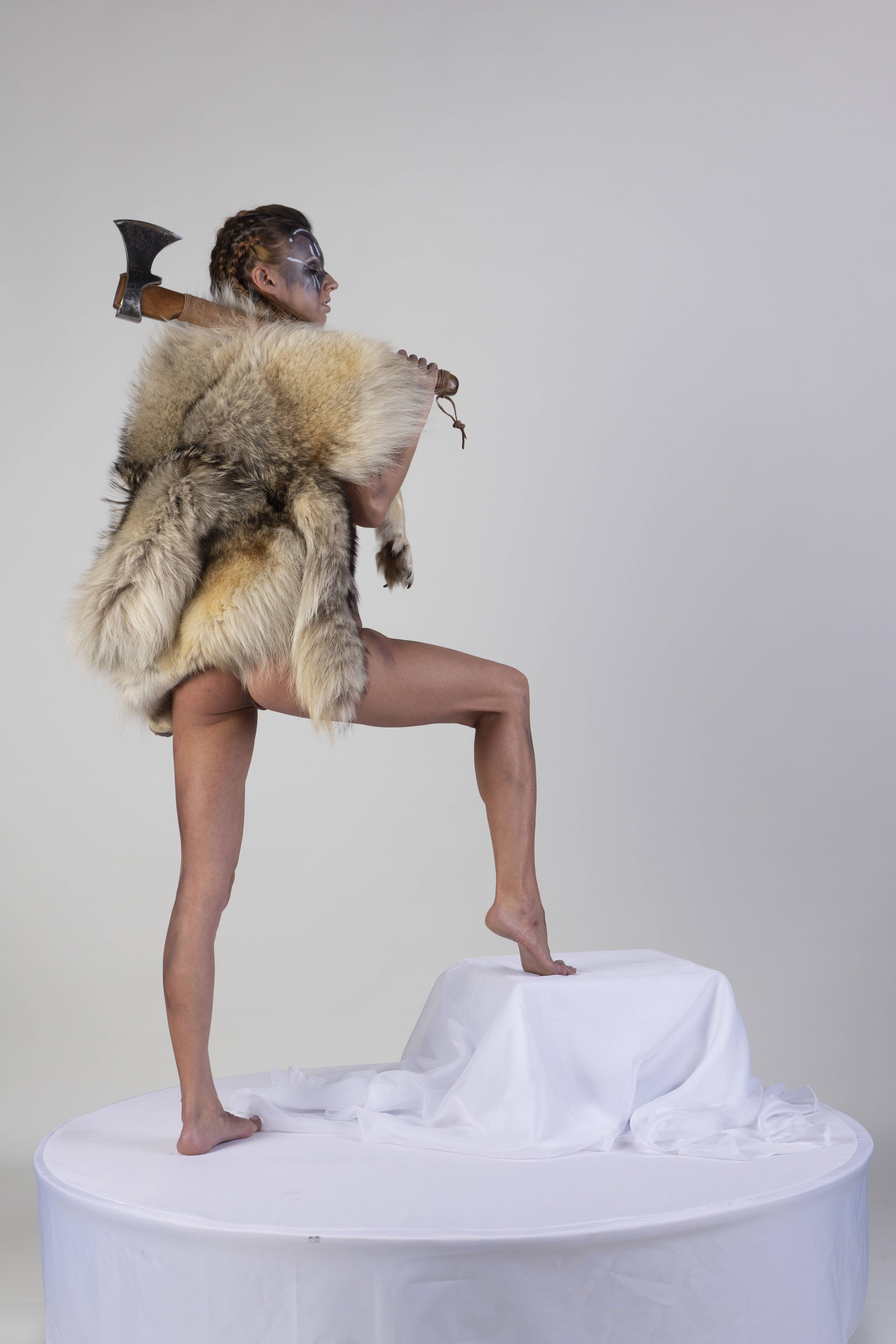 a woman in a fur covering stands on a white platform with an axe over her shoulder.