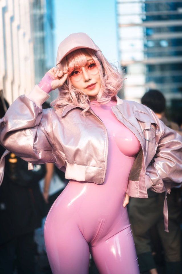 a young asian woman is striking a pose on a city street. she is dressed in a vibrant pink latex bodysuit and a metallic pink bomber jacket, giving off a futuristic and fashionable vibe. she has pink hair and is wearing a pink baseball cap and pink sunglasses, further enhancing the overall pink theme. the background suggests a bustling city environment, although the focus remains on the woman and her striking attire. the lighting is natural and sunny, casting soft shadows and highlighting the shine of the latex material.