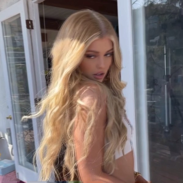 a woman with long blonde hair poses in front of a white door. the woman is wearing a white top and looks over her shoulder at the camera.